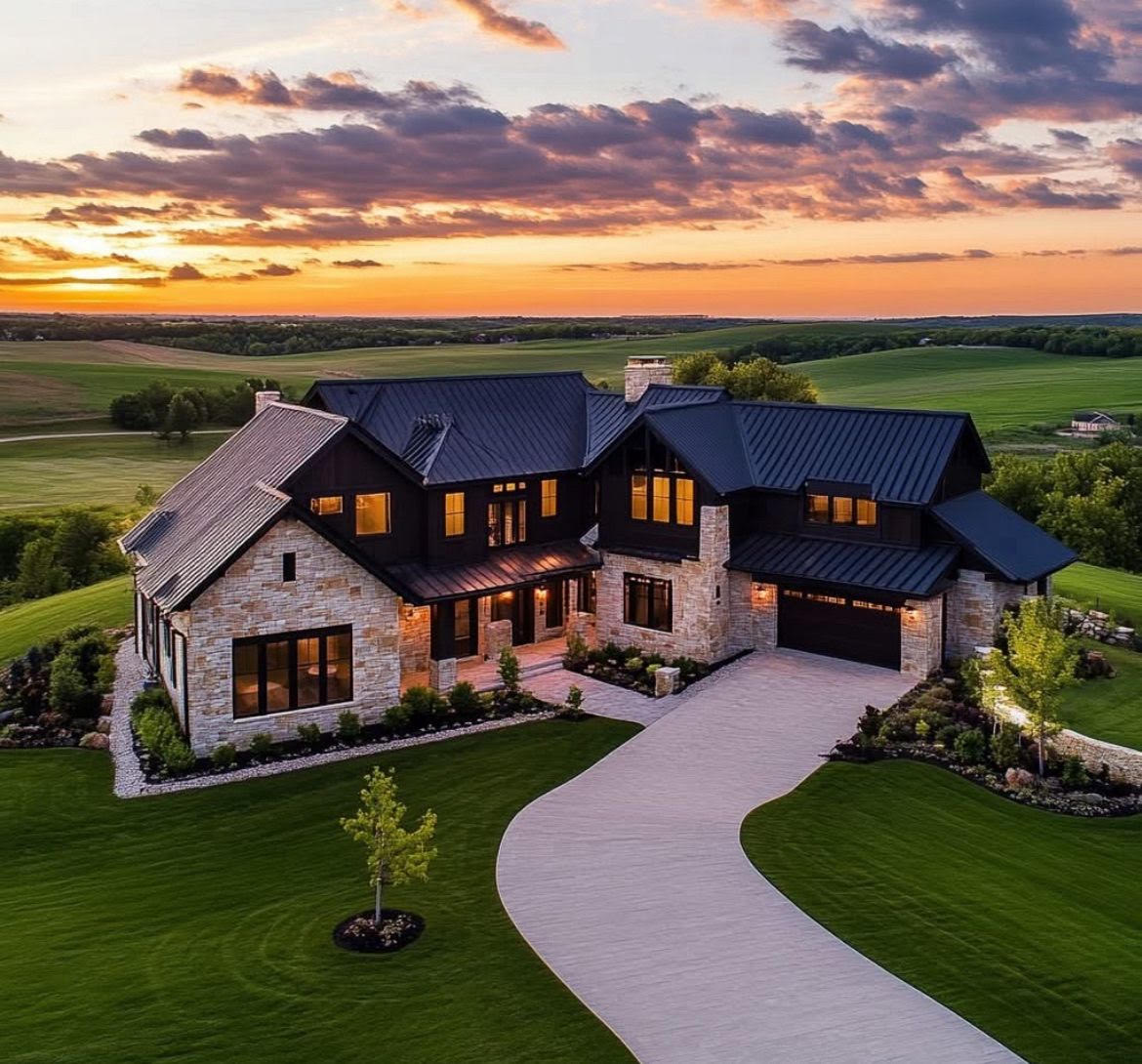 A stone and dark-sided estate home on a curving drive at sunset